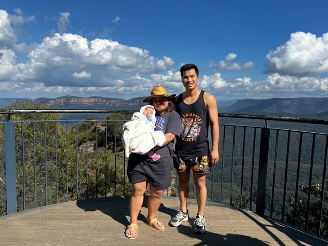 A man with his wife and baby standing together with a mountain view in the background. The man is wearing The Immortal Tiger tank top.